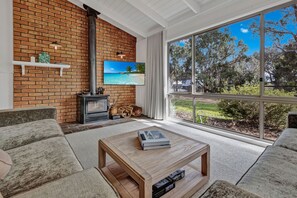 Un salon avec un mur en briques, une cheminée, un téléviseur et une grande fenêtre donnant sur les arbres et le ciel.