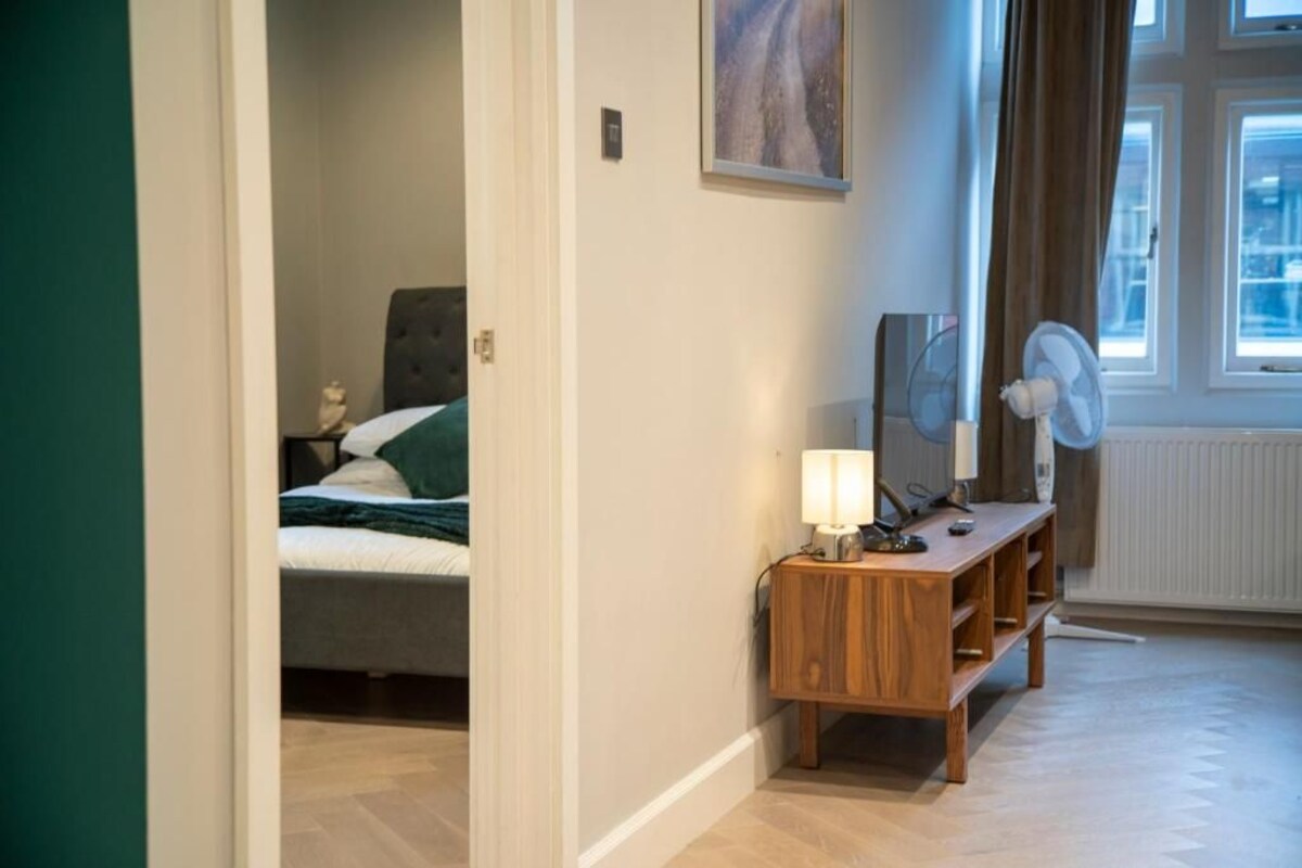 A bedroom with a bed, a bedside table with a lamp, a mirror, and a window with curtains.