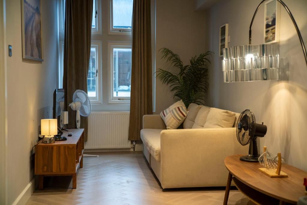 A cozy living room with a sofa, a wooden side table with a lamp, a fan, and a potted plant.