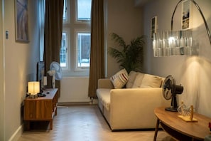 A cozy living room with a sofa, a wooden side table with a lamp, a fan, and a potted plant. - Berwick Soho Suites by Aureva Residences (London)