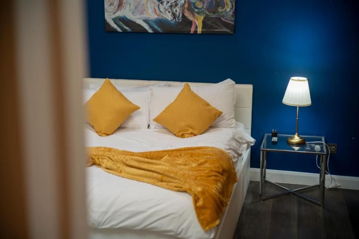 A bed with white bedding and two mustard pillows, a bedside table with a lamp, and a painting on the wall.