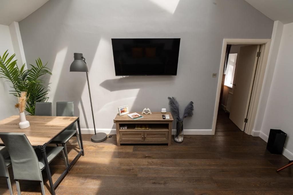 A modern living room with a flat-screen TV mounted on the wall, a wooden dining table with chairs, a floor lamp, and a small wooden cabinet.