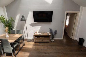 A modern living room with a flat-screen TV mounted on the wall, a wooden dining table with chairs, a floor lamp, and a small wooden cabinet. - Berwick Soho Suites by Aureva Residences (London)