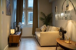 Una sala de estar con un sofá, una mesita, una lámpara, un ventilador y una ventana con cortinas.