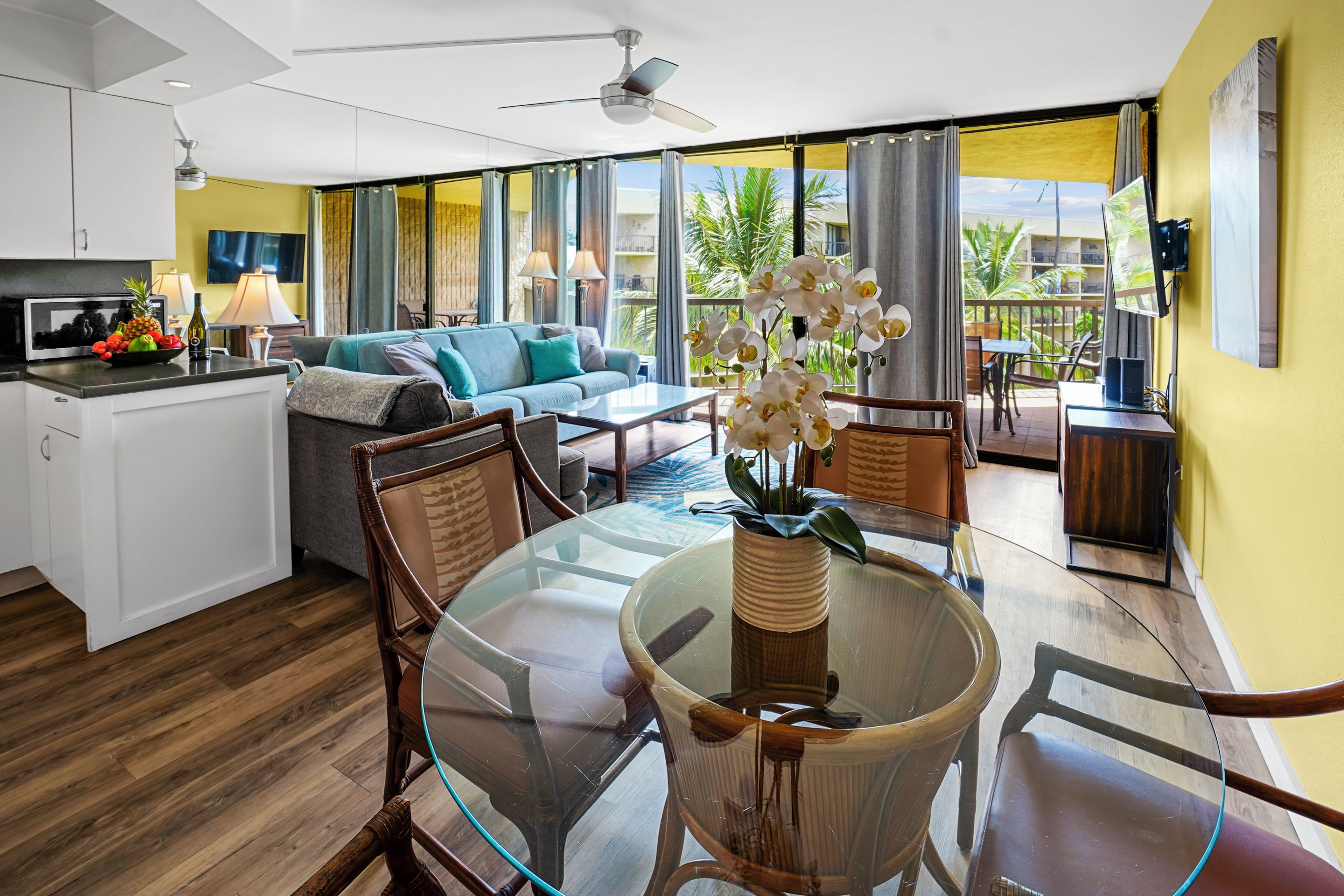 A modern living area with a glass dining table, wooden chairs, a sofa, and a kitchen with a microwave.