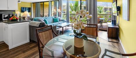 A modern living area with a glass dining table, wooden chairs, a sofa, and a kitchen with a microwave.