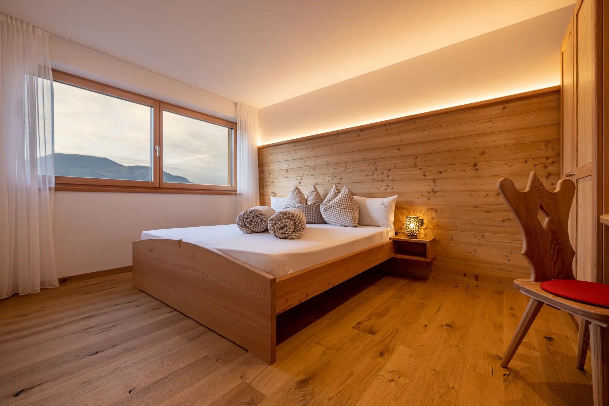 A bedroom with a wooden bed, a nightstand with a lamp, a chair, and a window with curtains.