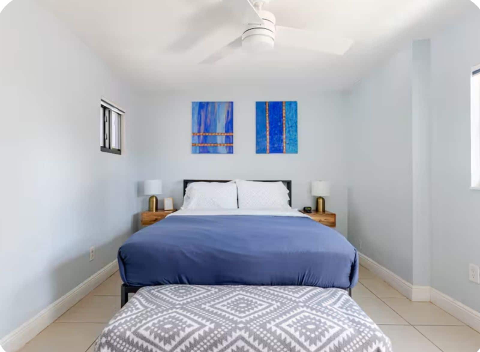 A bedroom with a bed, two nightstands, a ceiling fan, and two wall-mounted paintings.