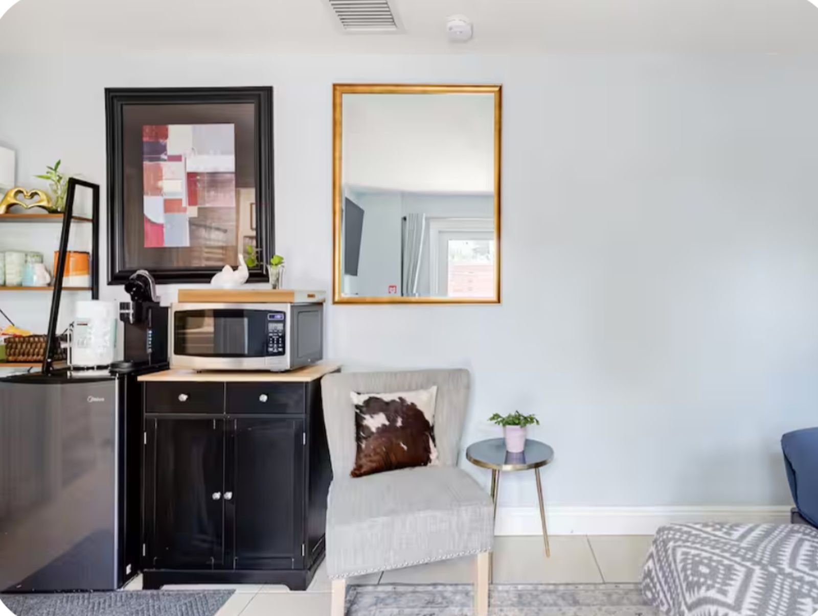 A compact living space with a kitchenette, a sofa, a small round table with a plant, and a large mirror.
