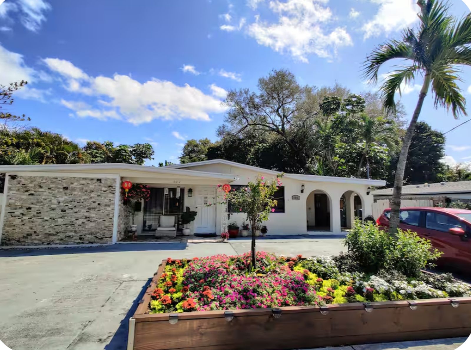 A two-story house with a front porch, a small garden with colorful flowers, and a palm tree.