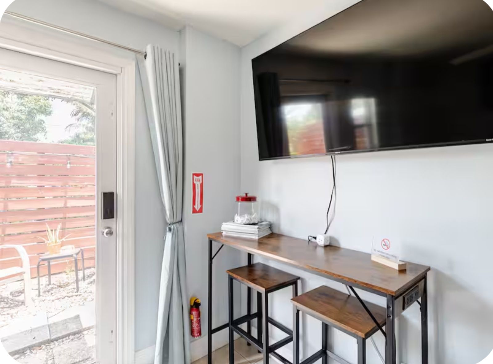 A room with a television mounted on the wall, a small table with stools, and a fire extinguisher on the wall.