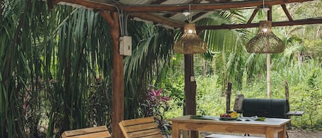 A covered outdoor seating area with wooden loungers, a table, and a grill.