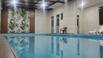 An indoor swimming pool with clear water, surrounded by walls with a tropical leaf pattern. There are two white lounge chairs and a red chair.