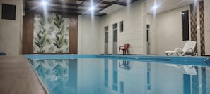 Une piscine intérieure à l’eau claire, entourée de murs ornés d’un motif de feuilles tropicales. On y trouve deux chaises longues blanches et une chaise rouge.