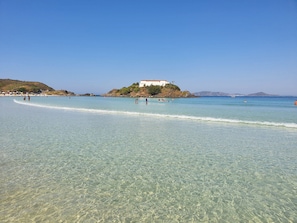 A beach with clear water, a white building on a hill, and people in the water. - Conforto e Elegância/1min.Praia do Forte (Cabo Frio)