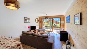 Ein Wohnzimmer mit einer Couch, einem Fernseher und einem Tisch mit einer Lampe. Ein Fenster bietet Blick auf einen Berg.