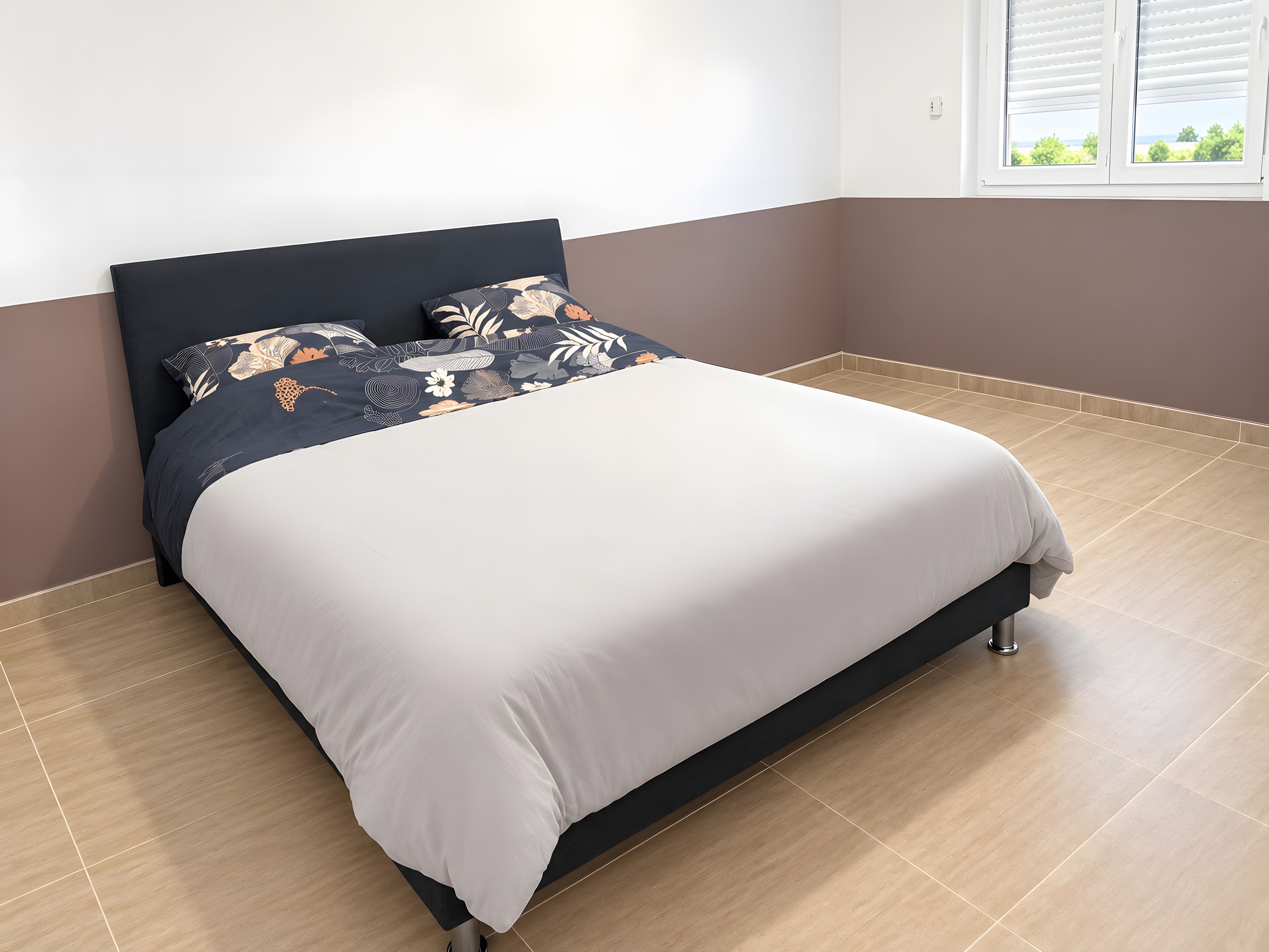 Un lit avec une tête de lit sombre et une couvre-lit à motifs floraux, un oreiller blanc uni et une couette de couleur claire.