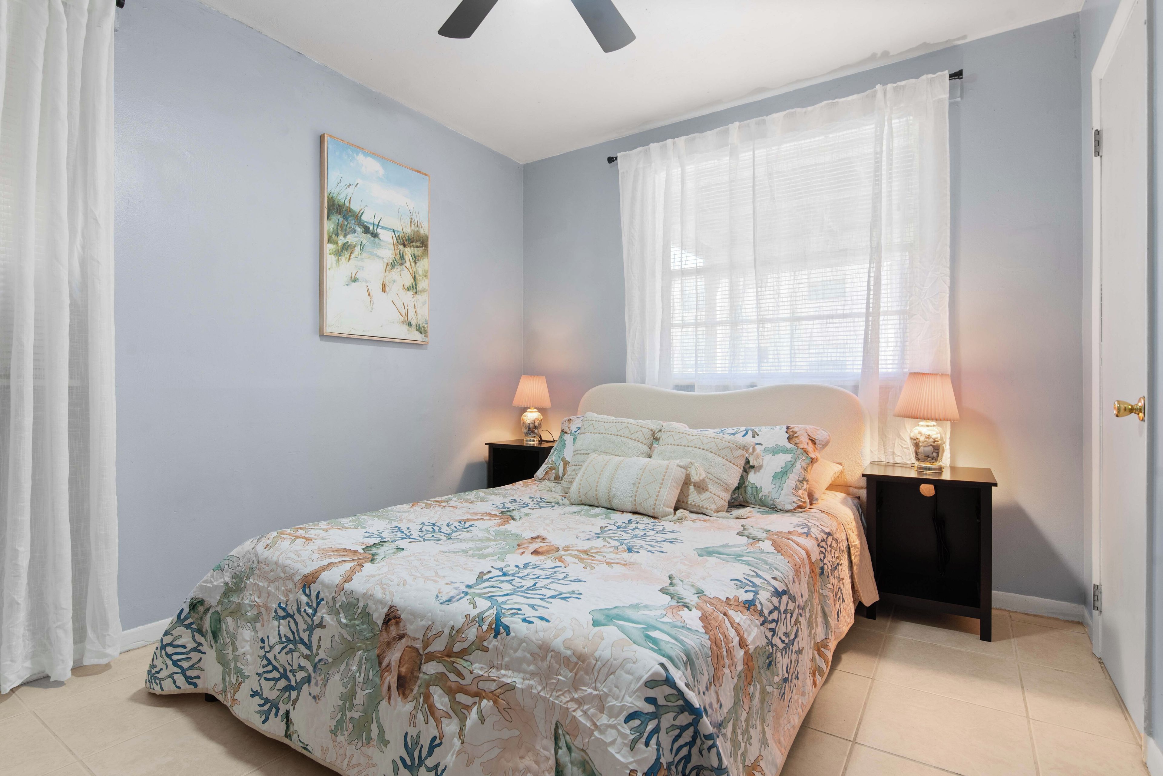 A bedroom with a bed, two bedside tables with lamps, a window with sheer curtains, and a painting on the wall.