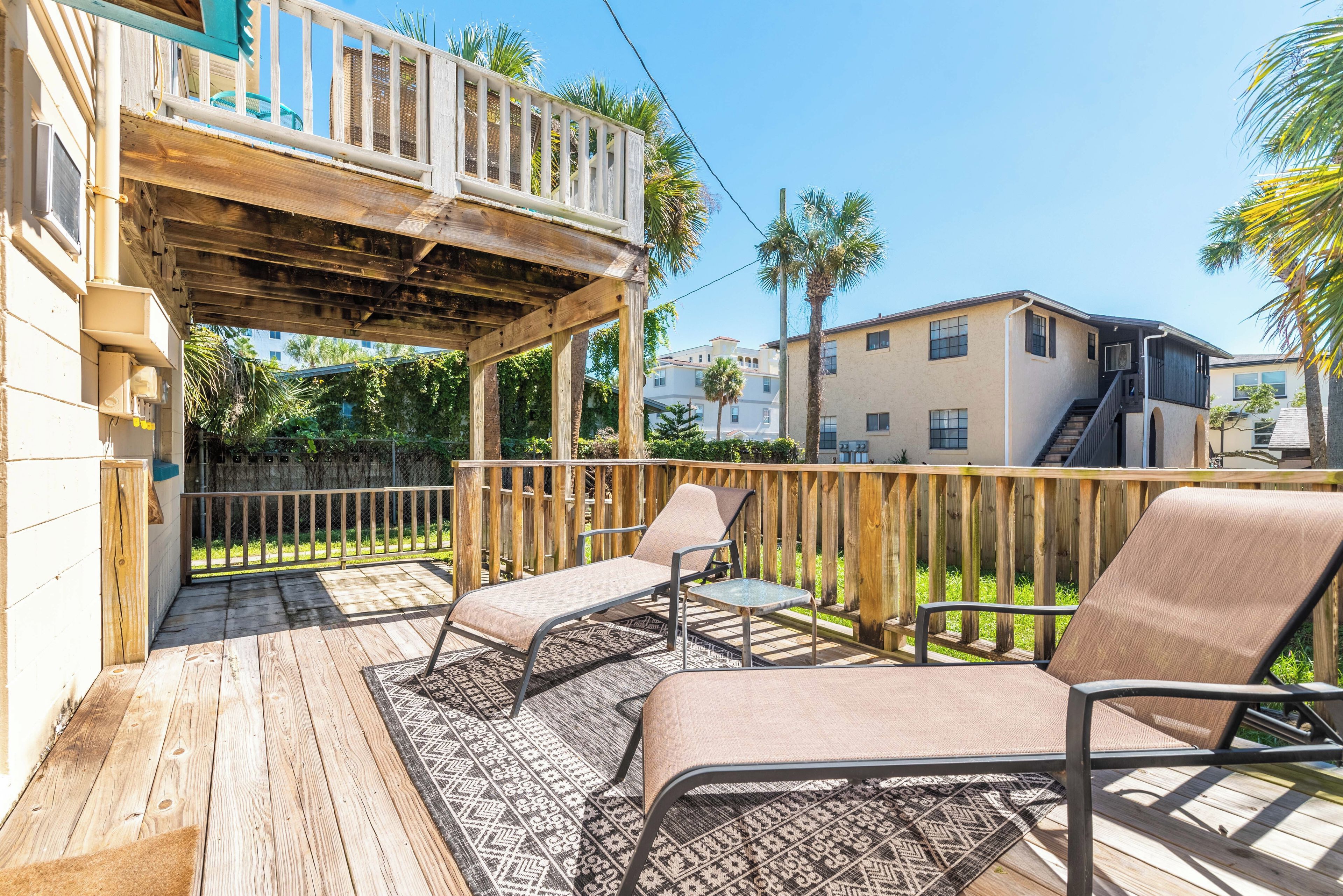 A wooden deck with two chairs and a patterned rug, overlooking a residential area with houses and palm trees.