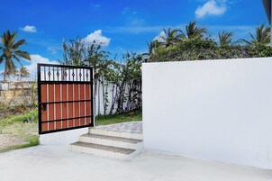 Uma entrada fechada com portão metálico, um caminho de concreto que leva a uma parede branca, e um céu azul claro com palmeiras ao fundo.