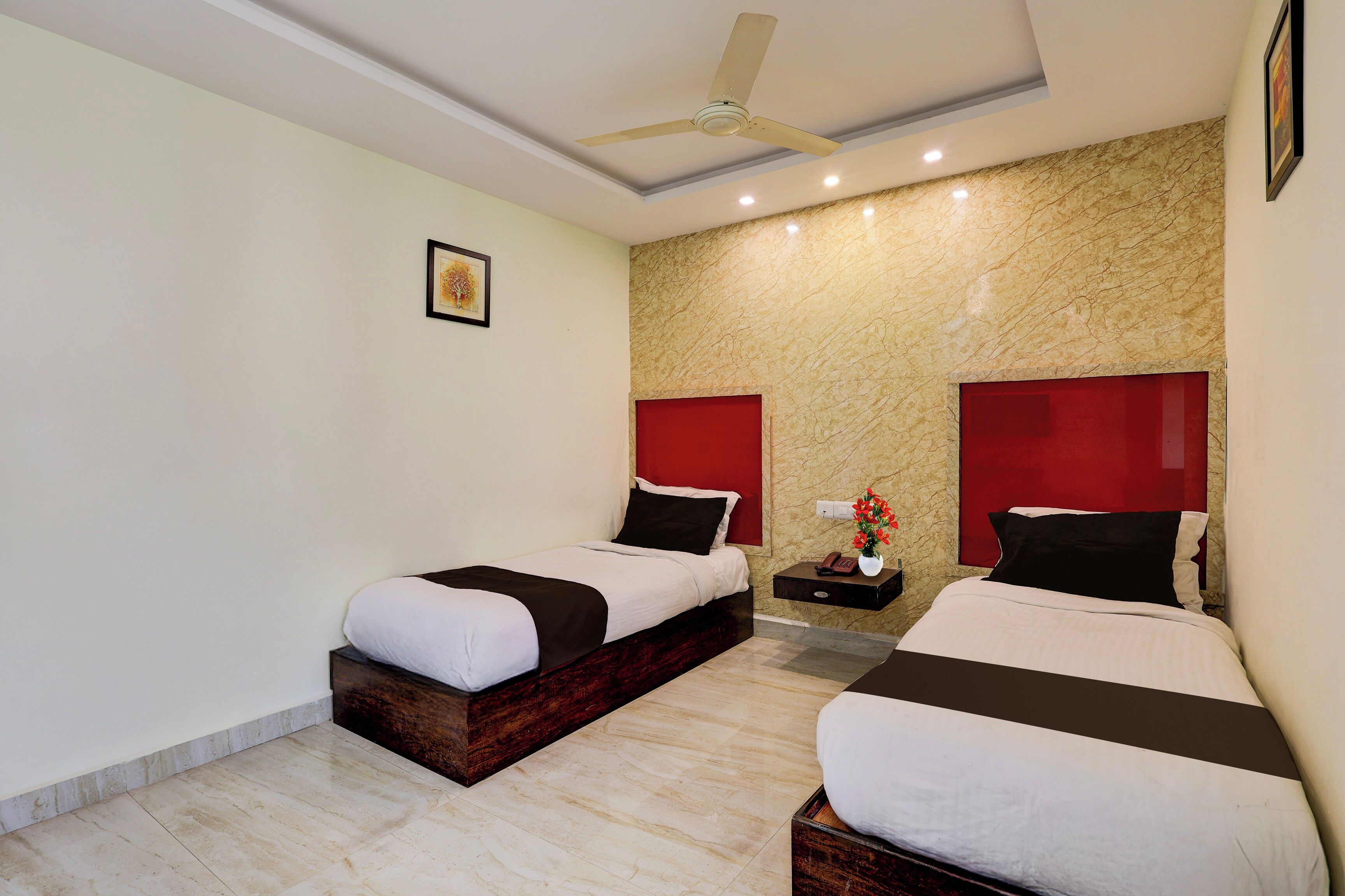 A hotel room with two beds, a ceiling fan, a marble wall, and a small bedside table with a vase of flowers.