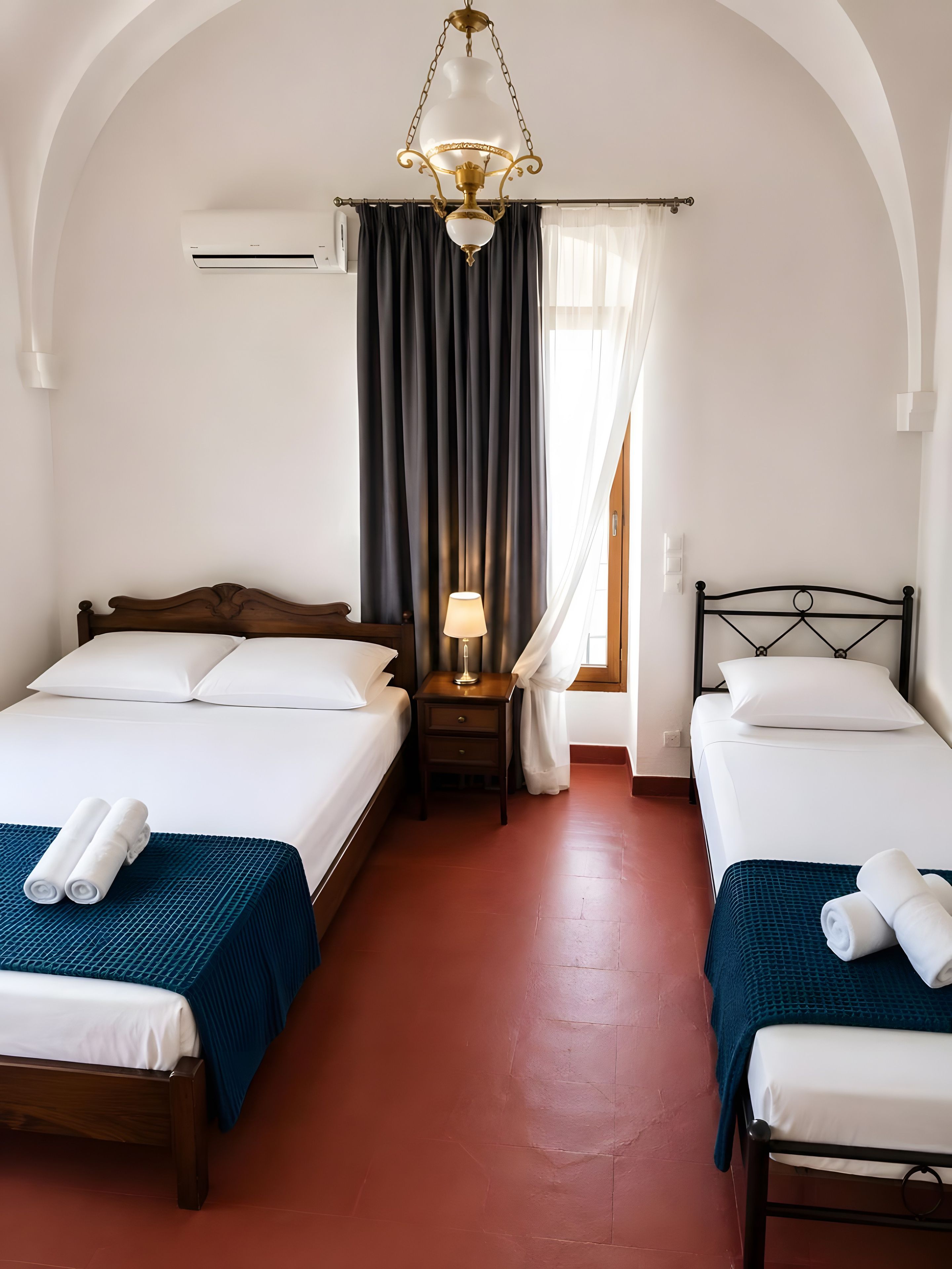 Quarto de hotel com duas camas, mesa de cabeceira com abajur, ar condicionado e janela com cortinas.