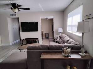 A modern living room with a sectional sofa, a flat-screen TV, a ceiling fan, and a window with blinds. - Gulf View Condo w/ 2 Kings | Steps to Beach + Pool (Fort Myers Beach)