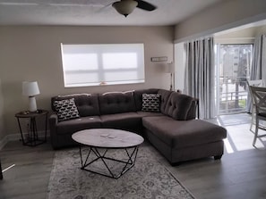A living room with a sectional sofa, a round coffee table, a lamp, and a view of the outdoors. - Gulf View Condo w/ 2 Kings | Steps to Beach + Pool (Fort Myers Beach)