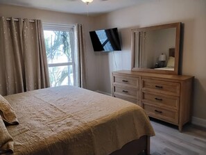 Ein Schlafzimmer mit einem Bett, einem Schminktisch, einem an der Wand befestigten Fernseher und einem Fenster mit Vorhängen.