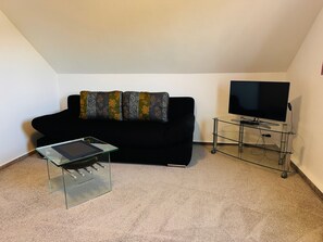 Une petite pièce meublée, avec un canapé noir, une table basse en verre, un téléviseur posé sur un meuble et un sol recouvert d’une moquette.