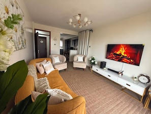 A living room with a sofa, armchairs, a TV displaying a fireplace, and a dining area in the background. - Nakuru View Palace Apartment (Nakuru)