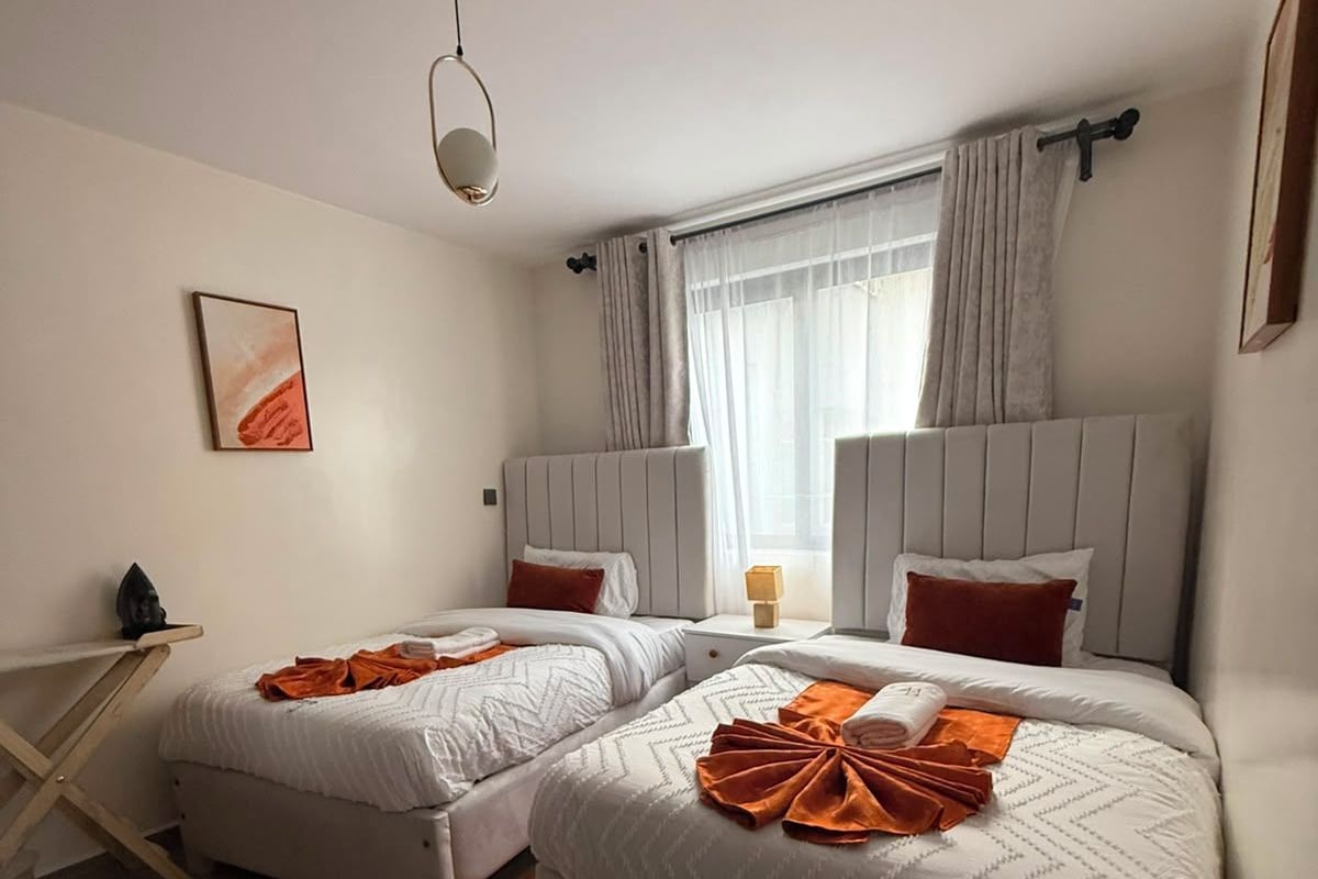 A hotel room with two beds, a bedside table, a small table, and a window with curtains.