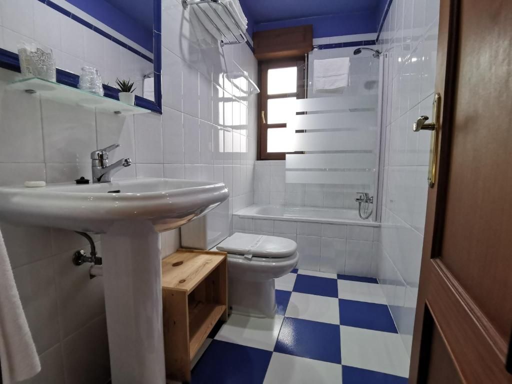 Un bany amb un rentapeus, un vàter i una banyera de color blanc, un paviment blau i blanc a quadres i una prestatgeria de fusta amb articles de tocador.