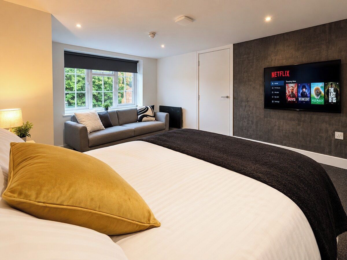 A modern hotel room with a large bed, a sofa, a TV mounted on the wall, and a window with a view of greenery.