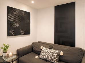 Una sala moderna con un sofá gris oscuro, un cojín estampado, una obra de arte abstracto en la pared y una mesita con una planta y una vela.