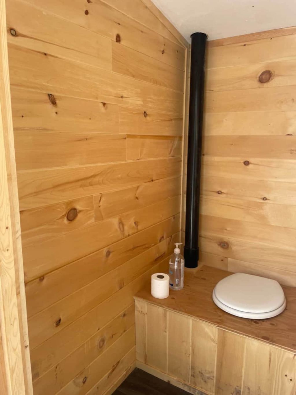 Une salle de bain rustique, aménagée dans une cabane en bois, avec des toilettes, un rouleau de papier toilette et un flacon de gel hydroalcoolique.