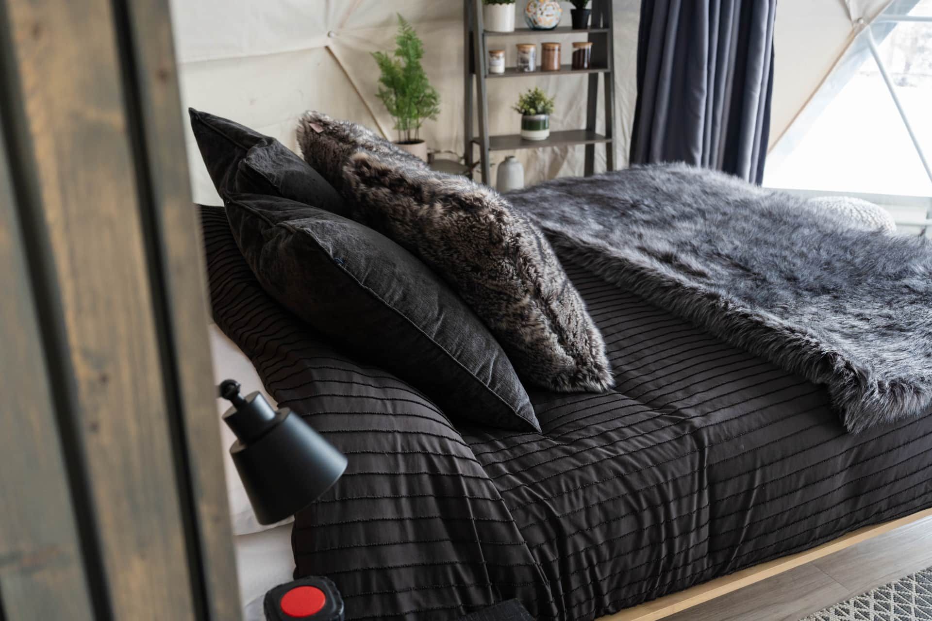 A bed with a dark quilted bedspread, multiple pillows, and a black lamp beside it.