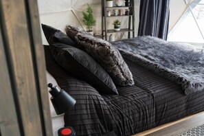A bed with a dark quilted bedspread, multiple pillows, and a black lamp beside it.