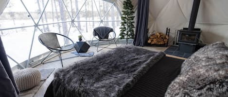 A tented room with a bed, two chairs, a small table, a Christmas tree, and a wood stove.