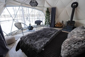 A tented room with a bed, two chairs, a small table, a Christmas tree, and a wood stove.