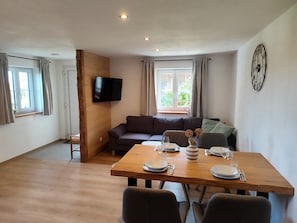 A dining area with a wooden table set for four, a sofa, and a television mounted on the wall. - Apartment 'Mair' with Private Terrace, Private Garden and Wi-Fi (Görisried)