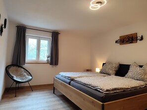 Un dormitorio con una cama de madera, una silla, un ventanal con cortinas, una lámpara de pared y un cuadro.