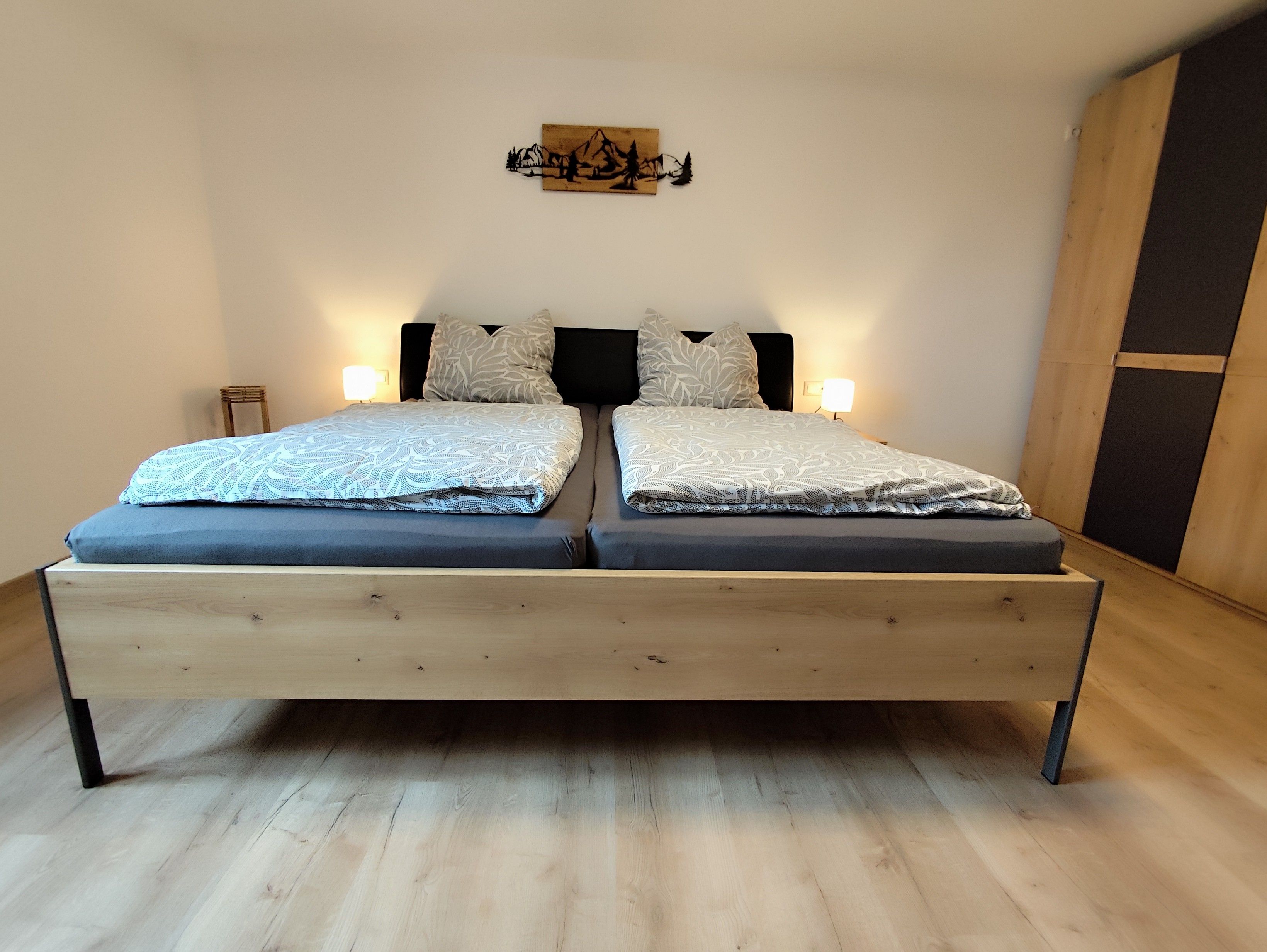 Ein Doppelbett mit Holzrahmen und gemusterten Betttextilien, zwei Nachttischlampen und ein an der Wand befestigtes Kunstwerk.
