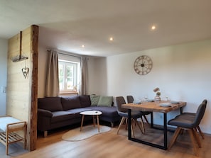 A modern living space with a sofa, dining table, chairs, a clock, and a window with curtains. - Apartment 'Mair' with Private Terrace, Private Garden and Wi-Fi (Görisried)