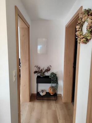 Un pasillo con un marco de puerta de madera, una consola negra con plantas y una pared blanca con un cuadro enmarcado.