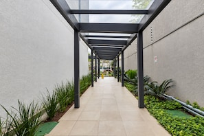 A covered walkway with a tiled floor, metal beams, and greenery on either side. - Studio in Vila Olímpia with Pool and Gym (São Paulo)