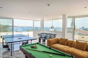 A pool table, foosball table, and a sofa in a room with large windows overlooking a body of water. - Villa Cecilia Coral - Six Bedroom Villa, Sleeps 12 (Peyia)