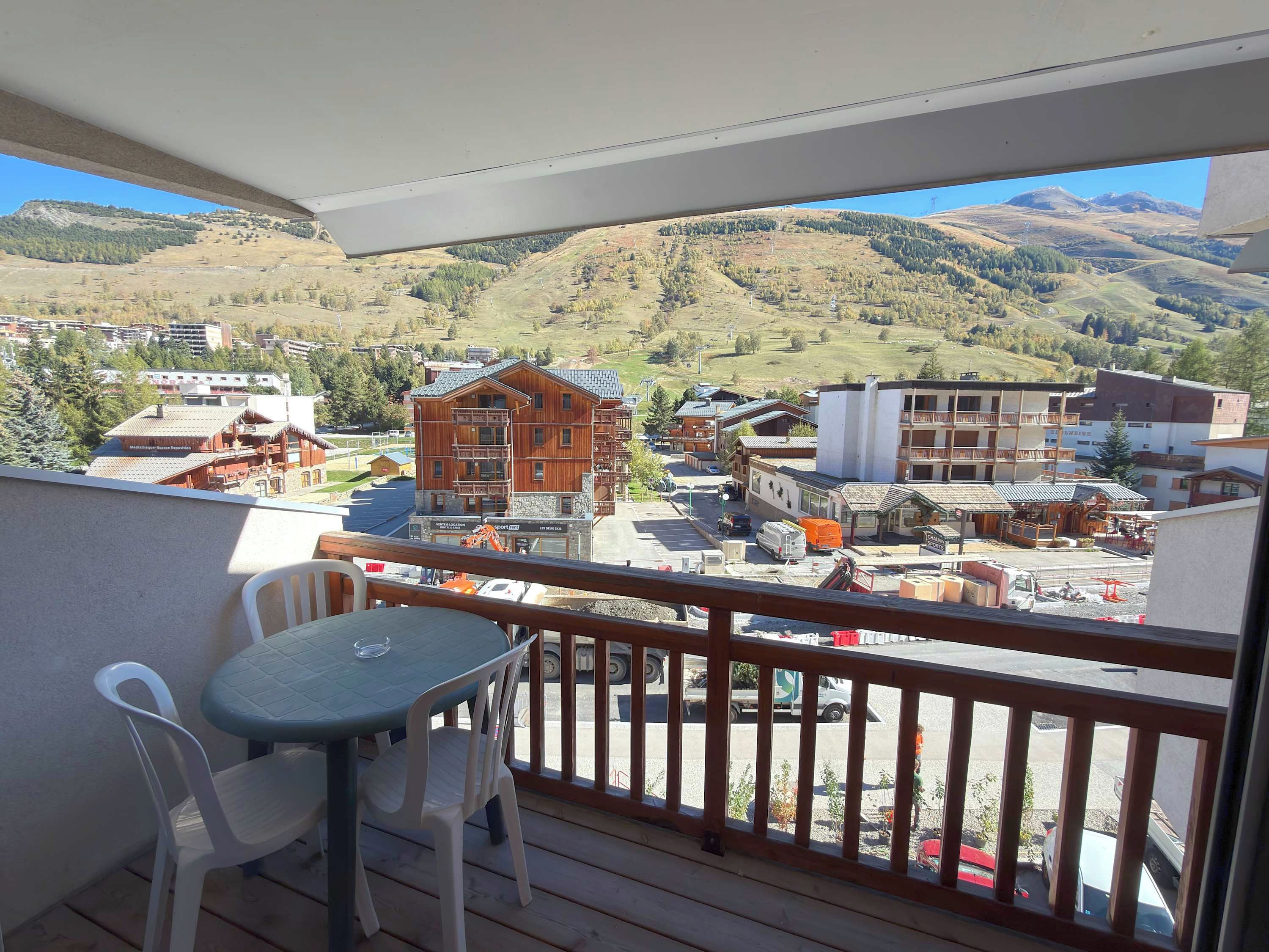 Un balcon avec une table et des chaises, donnant sur un paysage montagneux avec des bâtiments et un télésiège.