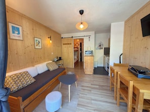 Ein gemütliches Zimmer mit Holzboden, einer Couch mit Kissen, einem kleinen runden Hocker, einem Esstisch mit Stühlen, einer Küchenzeile mit Mikrowelle und einem an der Wand befestigten Fernseher.