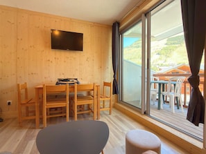 Ein Zimmer mit Holzvertäfelung, einem an der Wand montierten Flachbildfernseher, einem Esstisch mit Stühlen und einer großen Schiebeglasdoor, die zu einem Außenbereich mit Blick auf Häuser führt.